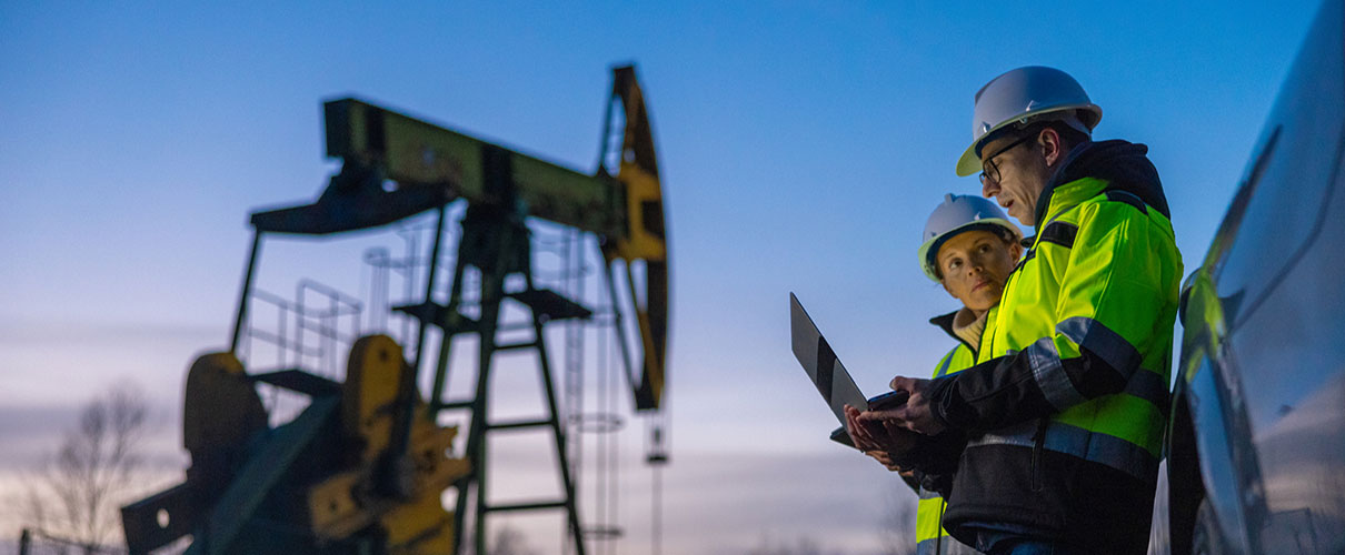 Two workers in high-vis jackets and hard hats look at a laptop while oil is drilled in the background
