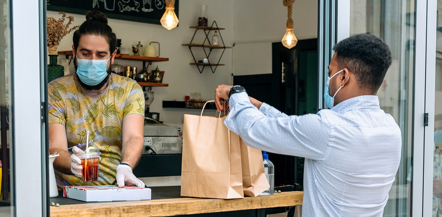 Masked restaurant owner handing order to masked customer through window