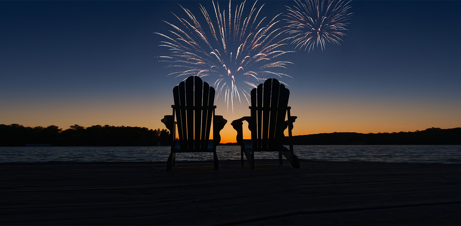 Two empty Adirondack chairs in front of a twilight fireworks display