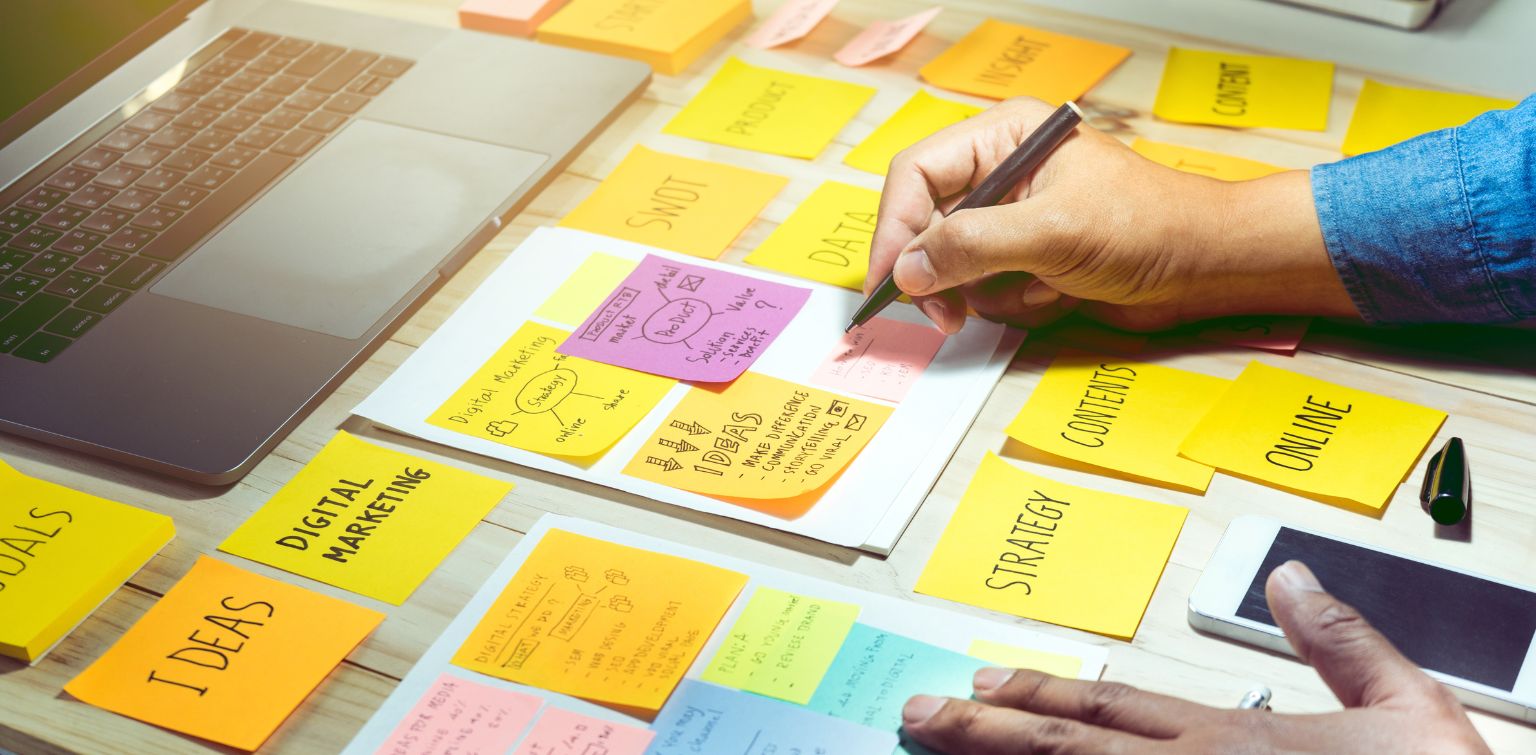 A laptop surrounded by colorful sticky notes across a desk, and hands writing more notes about marketing.