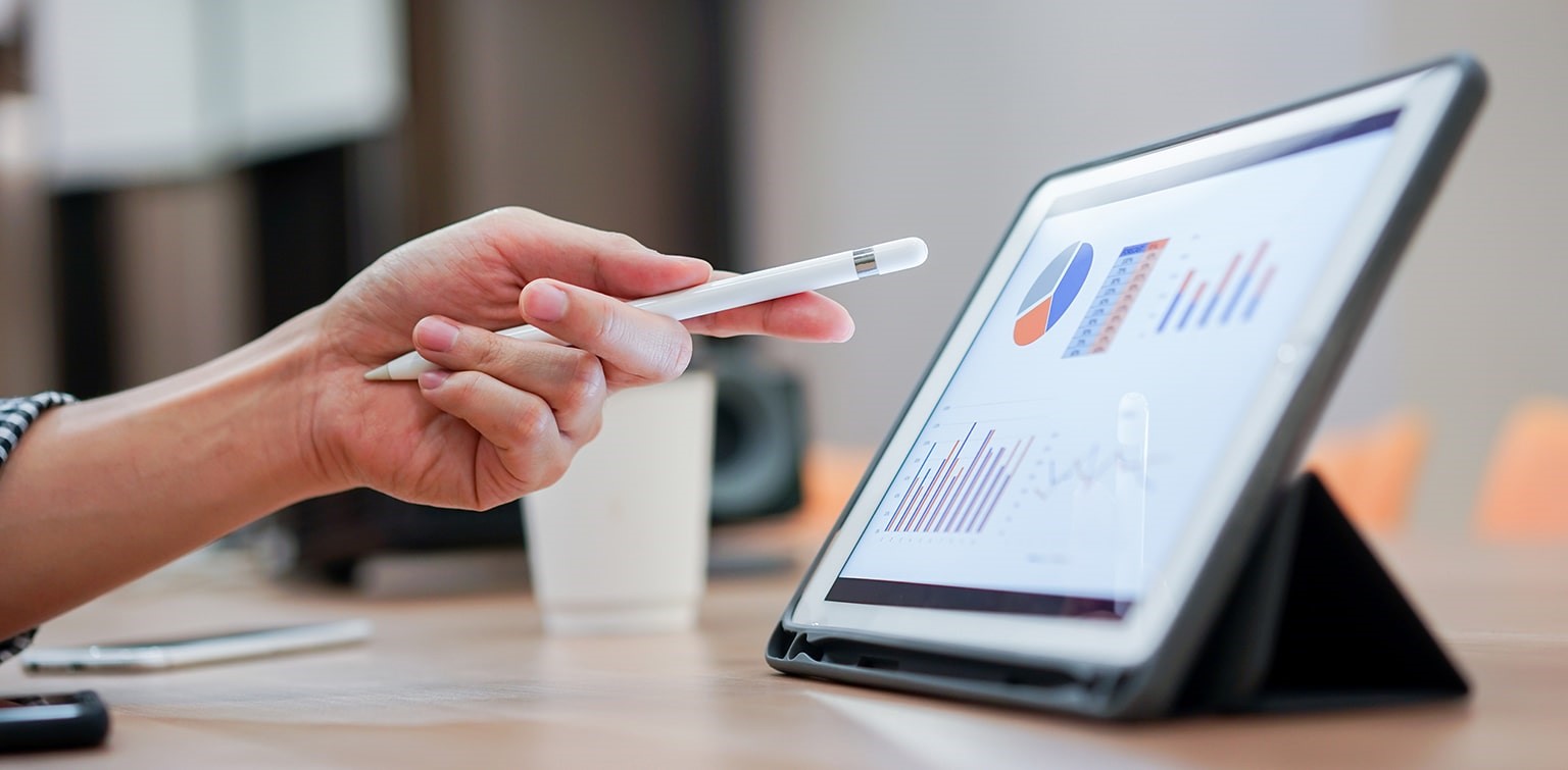 A hand pointing a stylus at a tablet screen displaying graphs and charts