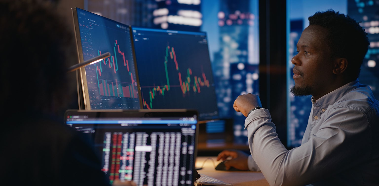 African-American financial analyst reviewing stock market data on two monitors