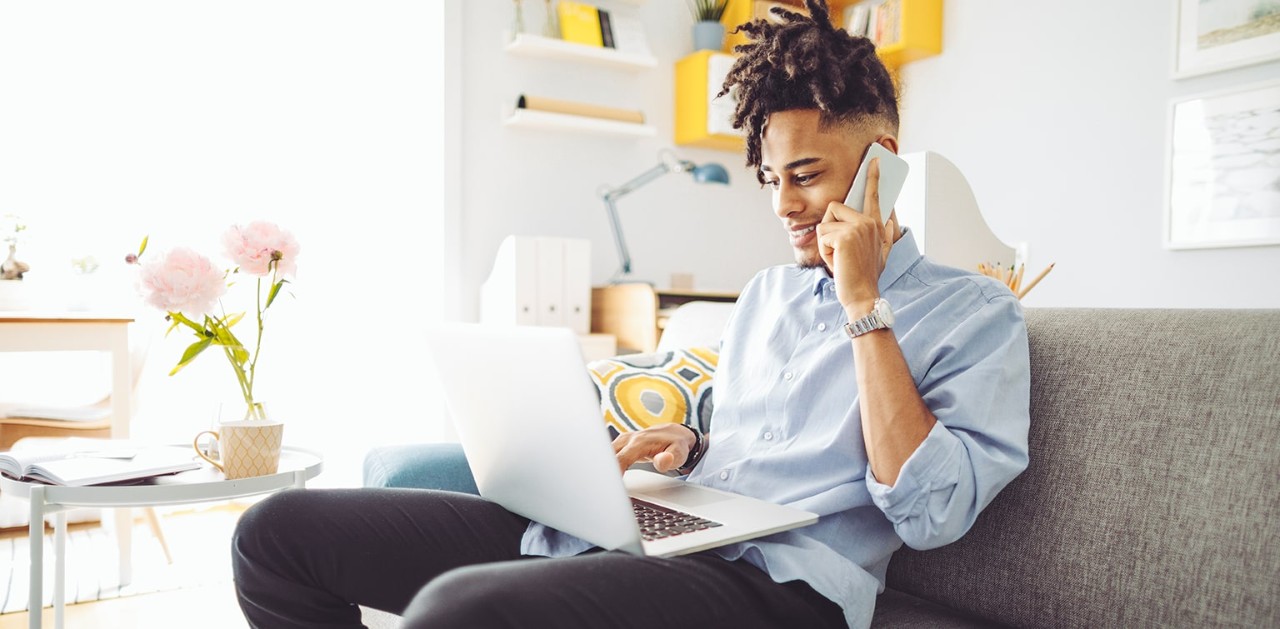 A person sitting on the couch talking on the phone while also typing on a laptop.