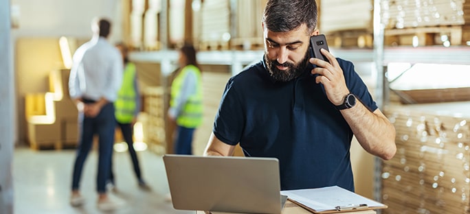 Man talking on smartphone and typing on laptop in a warehouse with coworkers in the background