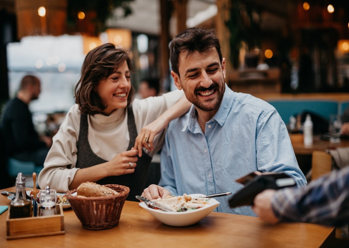 A couple sitting at a restaurant table enjoying a meal, with a basket of bread and a bowl of salad in front of them, as a server processes a payment with a card reader.