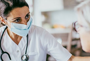 A medical professional in scrubs in mask with a stethoscope around her neck observing a patient.