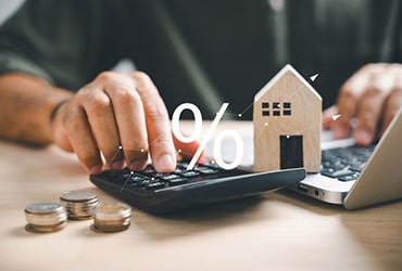 Close-up of a man's hands using a calculator and laptop, with coins and wooden house model nearby