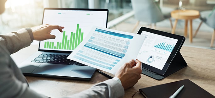 Analyst reviewing financial data on a laptop, tablet, and paper documents