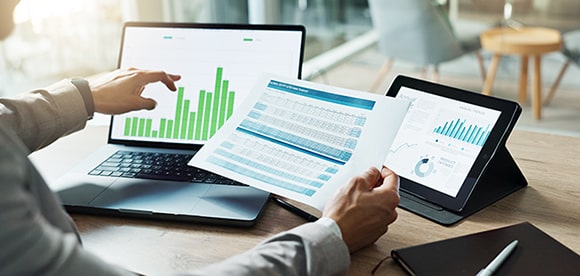Analyst reviewing financial data on a laptop, tablet, and paper documents